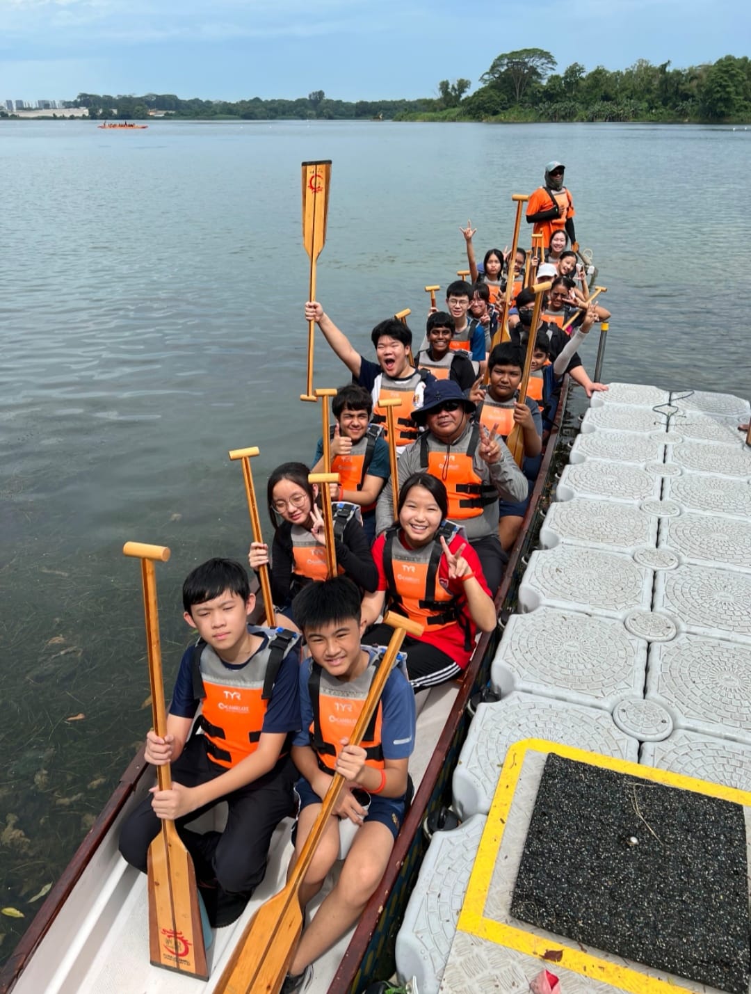 A group of young people wearing orange life vests sit in a long dragon boat on a river, holding wooden paddles and smiling.